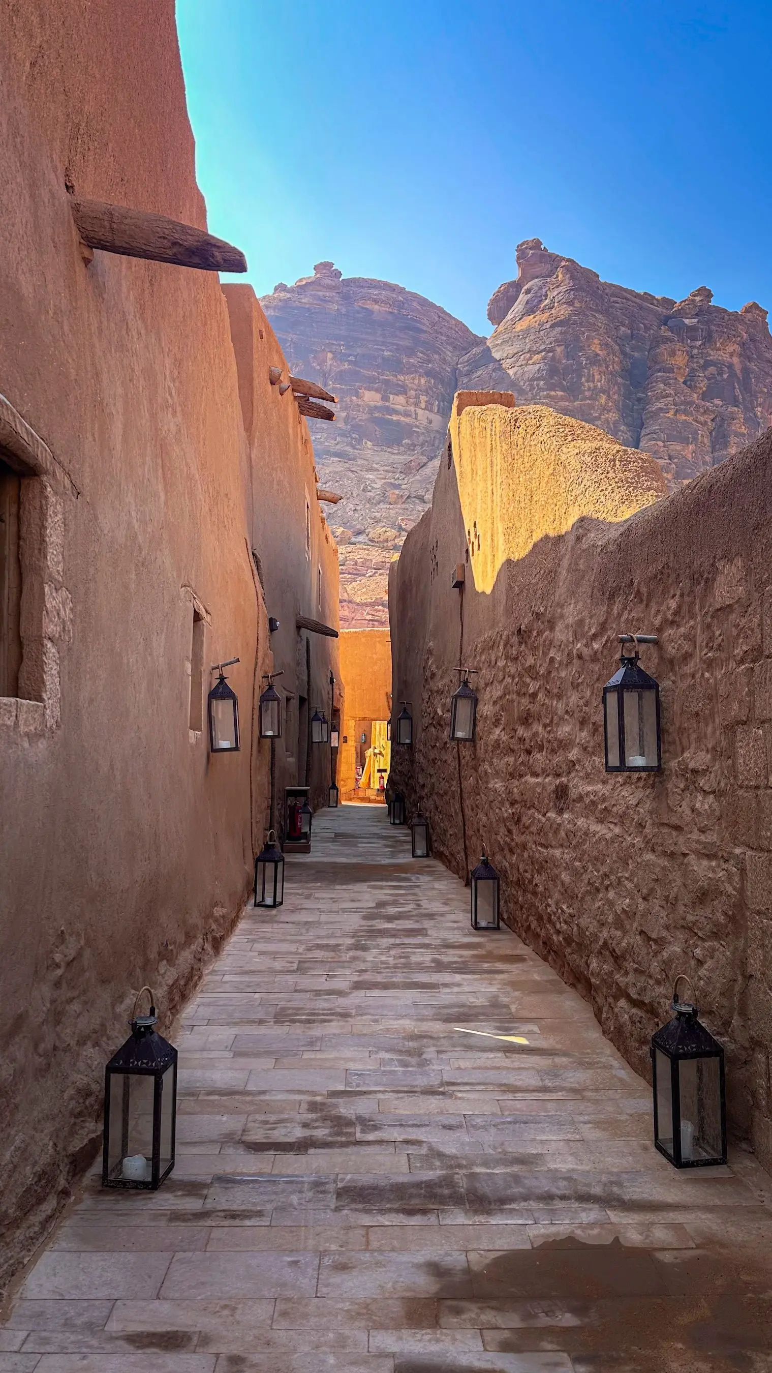 A narrow earthen alley in AlUla with desert cliffs rising behind the houses.