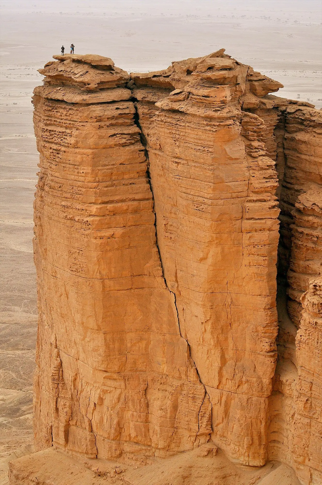 Two small figures stand on a narrow sandstone pinnacle above a desert plain.