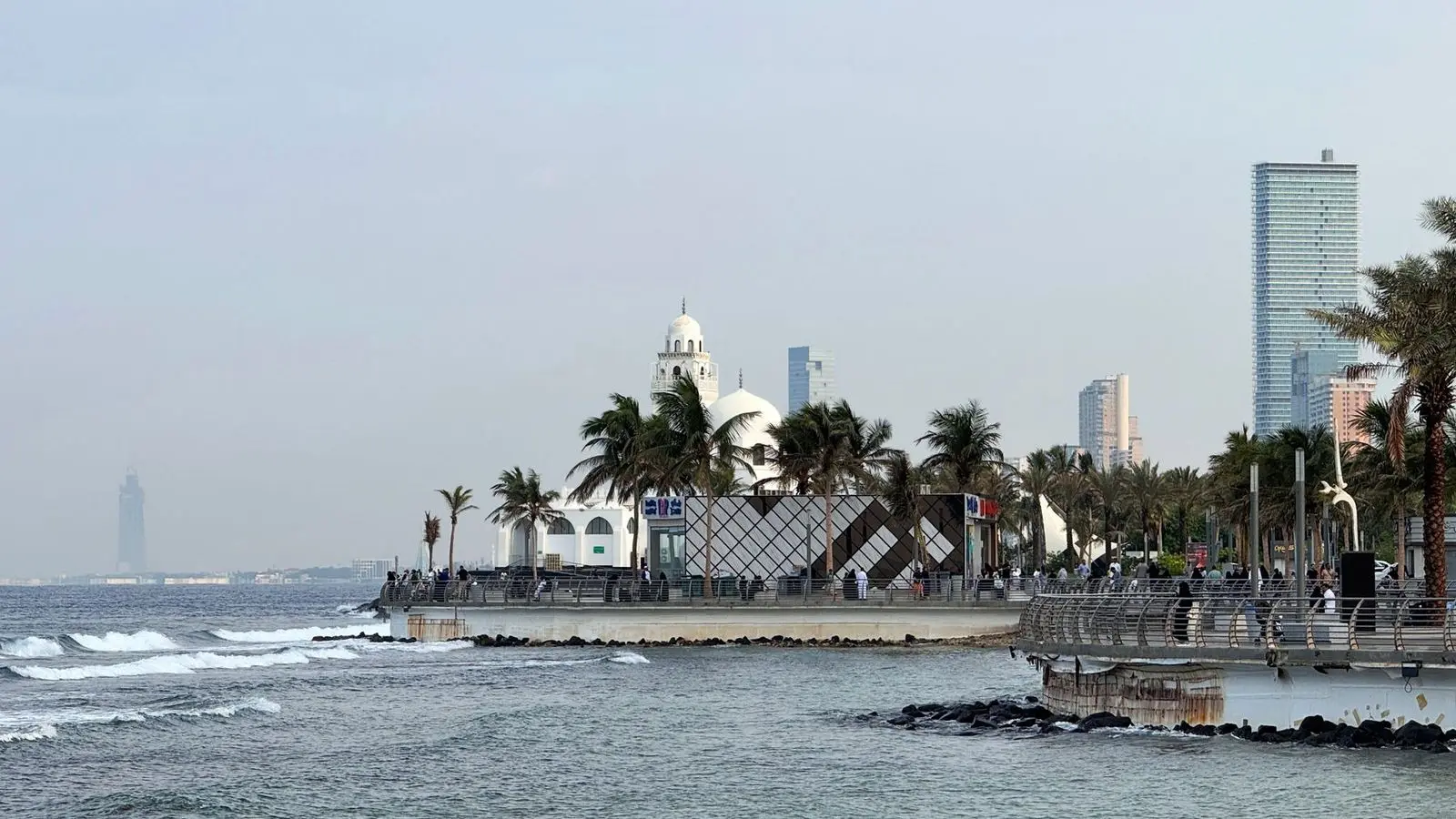 A white mosque and palm trees along a paved seafront, with waves breaking on rocks and towers behind.